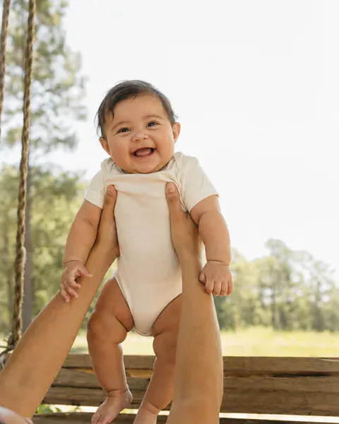 A smiling baby held up by a parent’s arms under the pines.